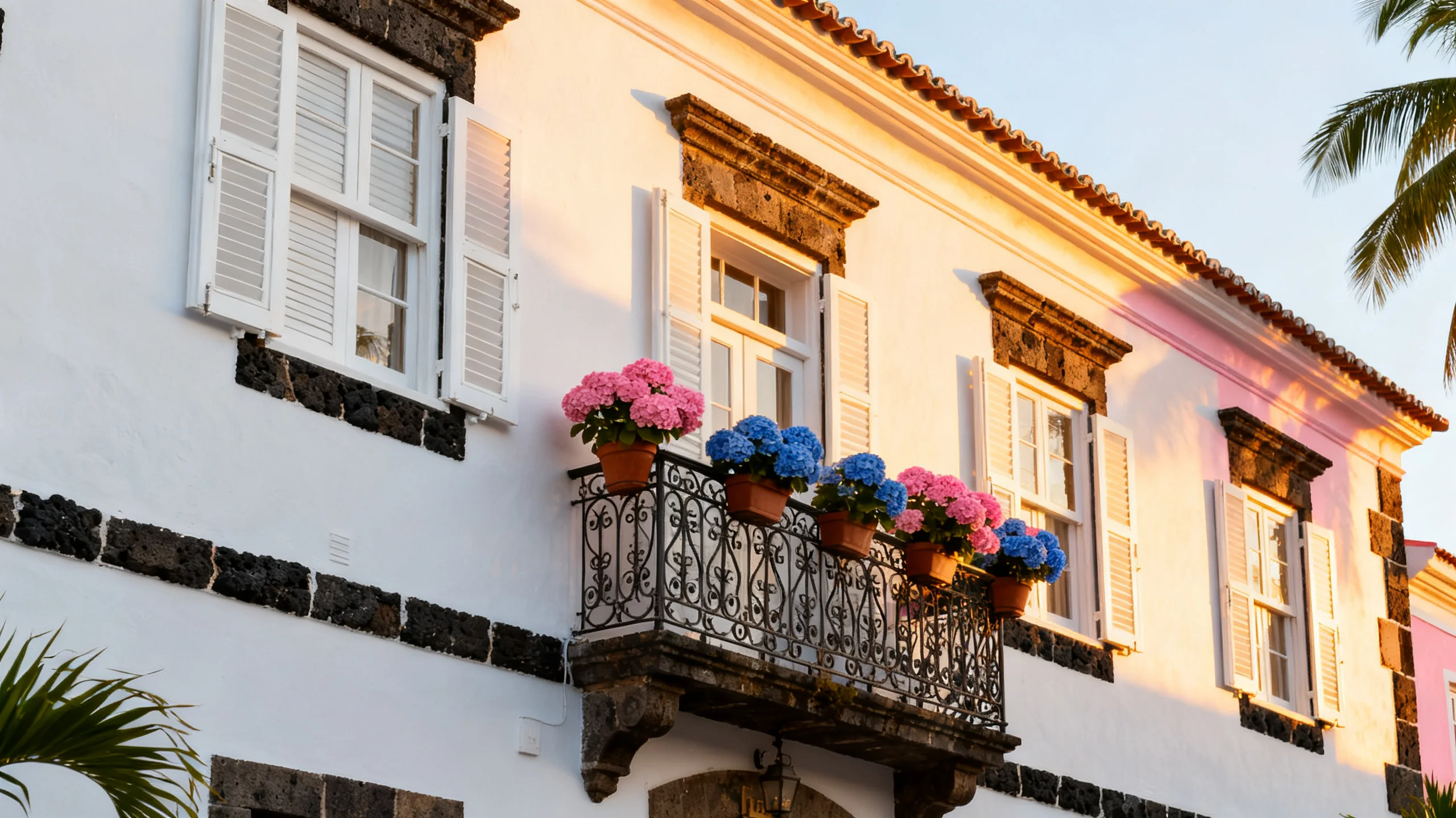 A boutique hotel in Ponta Delgada, São Miguel, with a restored 19th-century facade in white and pastel paint, a wrought-iron balcony with hydrangea flower pots, the warm afternoon golden light hitting the white walls