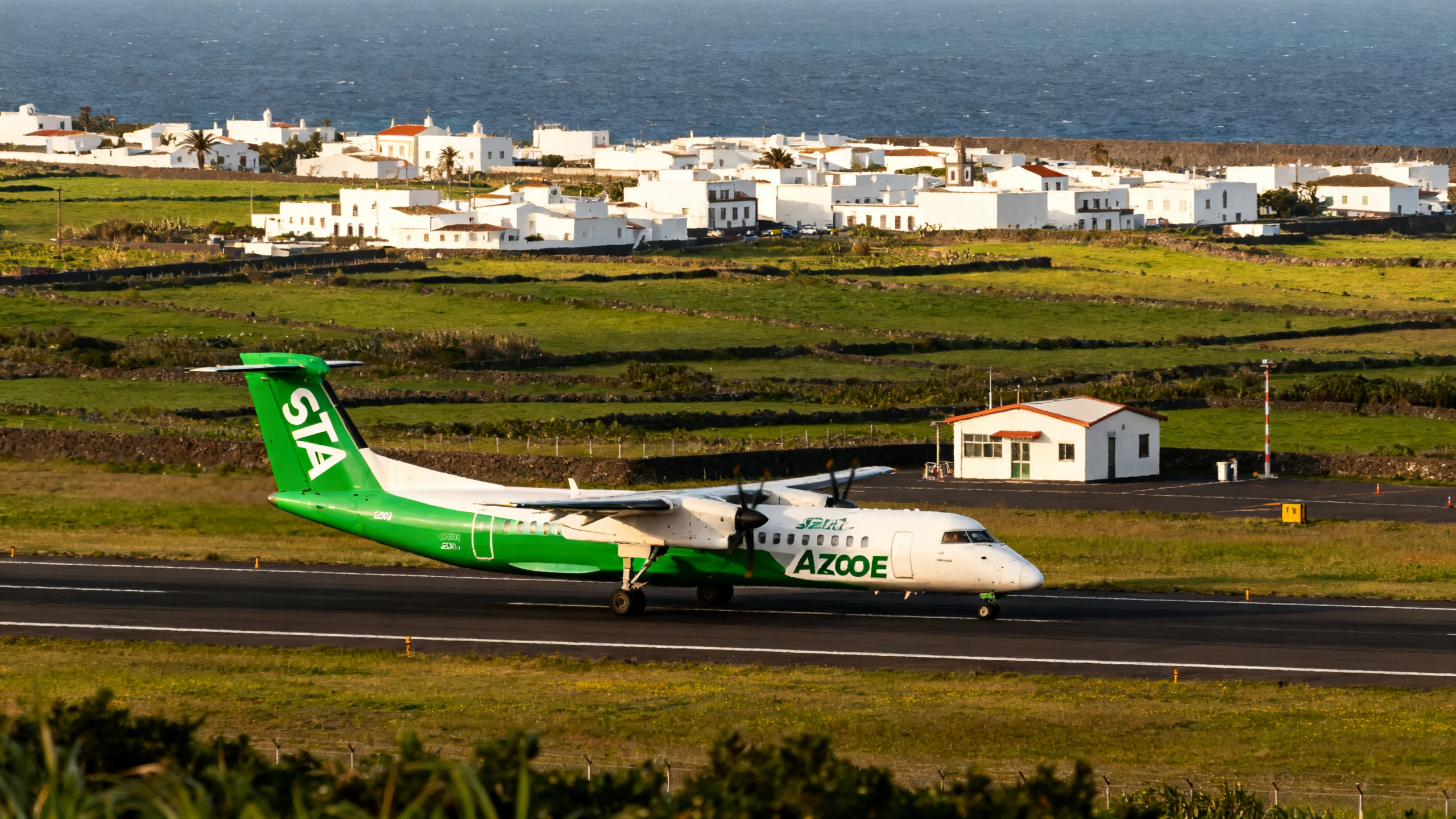 A SATA Azores Airlines Bombardier Q200 turboprop on the small runway of Graciosa Airport, the white-painted town of Santa Cruz da Graciosa and the surrounding green pastoral landscape visible in the background under soft afternoon light, the small terminal building visible at the edge of the runway