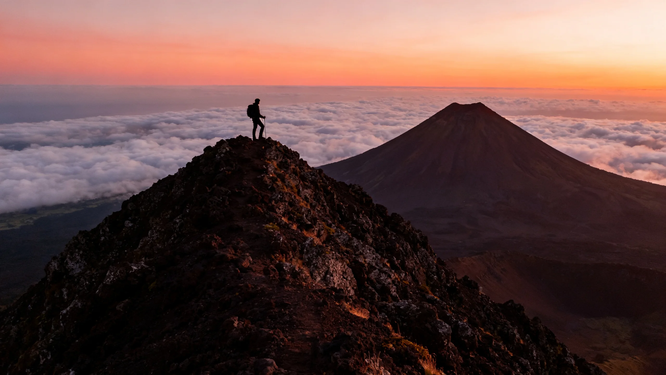 A hiker silhouetted on the summit ridge of Pico volcano in the Azores, with the small summit cone Piquinho rising in the foreground, a sea of clouds covering the lower slopes far below, and the orange and pink colours of dawn lighting the volcanic rock