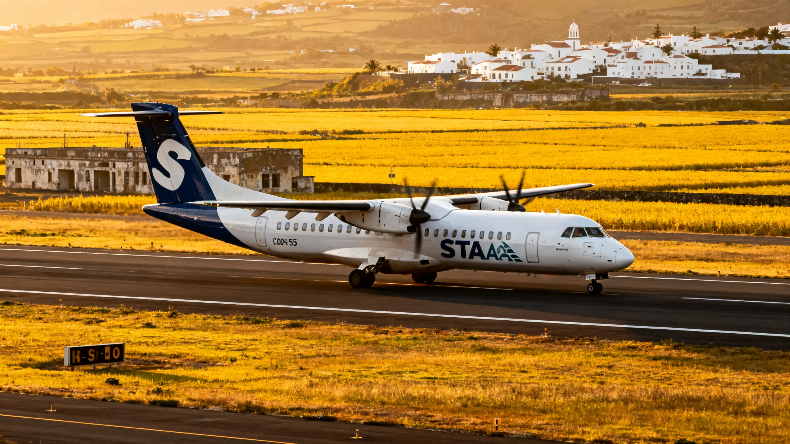 A SATA Azores Airlines aircraft on the long runway of Santa Maria Airport, the legacy of the WWII US military base, with the yellow-tinted agricultural landscape of the island visible in the background, the village of Vila do Porto in the distance, soft afternoon golden light
