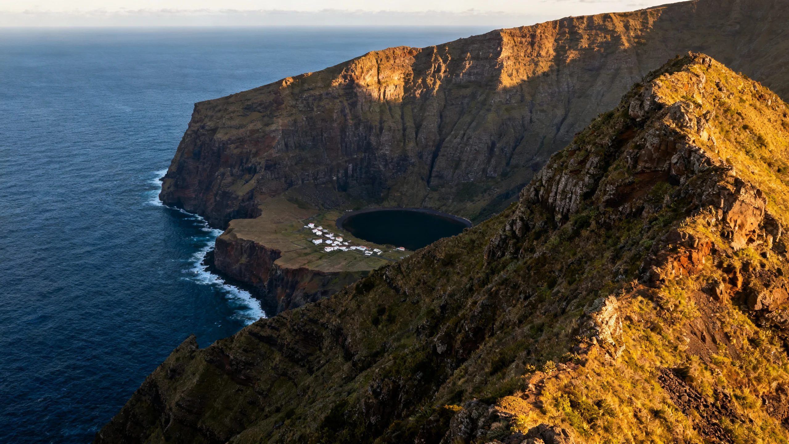 A panoramic view down from the ridge of São Jorge into Fajã da Caldeira de Santo Cristo, showing the flat coastal platform with its lagoon, a small village of white-painted houses, the towering basalt cliff backing the fajã, and the deep blue Atlantic ocean to the horizon under late-afternoon golden light